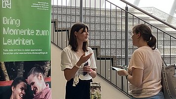 Karrierestand mit zwei Frauen im Gespräch, zwischen ihnen ein Tisch mit weißer Tischdecke. Darauf liegen grüne Flyer, ein aufgeschlagenes Notebook, Stiftebecher, rosa Post-Ist und ein hochkant aufgestellter Prospektständer. Links hinter dem Tisch steht ein großes grünes Rollup. Darauf oben ein weißes Logo mit stilisiertem „N“ und der Text „Nikolauspflege Stiftung für blinde und sehbehinderte Menschen“. Darunter in großen weißen Buchstaben „Bring Momente zum Leuchten. Im Hintergrund hinter der Szene führt eine Treppe schräg nach links oben.