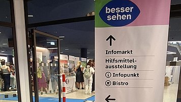 Roll-up-Banner in einer Messehalle. Rechts im Bild steht ein hohes, schmales Banner mit weißer Fläche und farbigem Kopfbereich, links davon verläuft ein breiter, kräftig blauer Wegstreifen über den Boden in einen offenen Bereich mit mehreren Personen und Ständen. Oben auf dem Banner steht: „besser sehen“ Darunter folgen Pfeile und Text: Nach oben gerichteter Pfeil „Infomarkt. Hilfsmittelausstellung. Infopunkt. Bistro“. Darunter ein nach links gerichteter Pfeil „Raum 10–25. Aktionsfläche. Berliner Platz. Aufzug. WC“