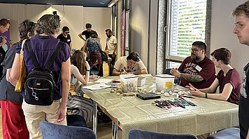 Ein Gruppenraum mit mehreren Menschen an einem langen Tisch beim Basteln und Malen. Der Raum wirkt hell, mit großen Fenstern rechts und hellem Holzboden.   In der Mitte steht ein langer rechteckiger Tisch mit hellbeiger Tischdecke, darum herum stehen blaue gepolsterte Stühle mit Metallbeinen. Auf dem Tisch liegen viele bunte Stifte, Filzstifte und andere Malutensilien, außerdem verschiedene Schachteln, Becher und kleinere Gegenstände, die wie Bastelmaterial wirken. Rund um den Tisch sitzen und stehen Menschen unterschiedlichen Alters, die meisten tragen Freizeitkleidung wie T Shirts, Hosen und Rucksäcke. Im Vordergrund steht eine Person mit kurz gelockten, dunklen Haaren, lila Oberteil, heller Hose und schwarzem Rucksack, die zum Tisch gewandt ist. Links hinten im Raum stehen mehrere Personen zusammen und unterhalten sich, einige tragen Rucksäcke oder Umhängetaschen. Rechts am Tisch sitzen zwei Personen nebeneinander; eine trägt ein dunkelrotes T Shirt mit weißem Aufdruck und Namensschild, die andere hat kurzgeschnittene Haare und ein dunkles Oberteil. Rechts am Bildrand ist der Oberkörper einer weiteren Person in einem T Shirt zu erkennen. Die Decke ist dunkelblau mit länglichen Lampen, die Wände sind hell, und durch die Fenster mit Jalousien dringt Tageslicht.