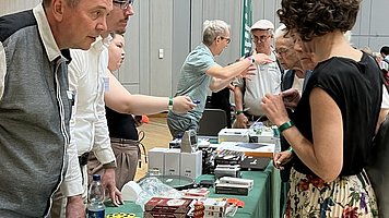 Messestand mit verschiedenen Hilfsmitteln auf einem grünen Tisch.   Auf dem Tisch liegen viele kleine Produkte ordentlich ausgebreitet: vorne eine leuchtend gelbe Tasche mit Reflexstreifen und einem dunklen, runden Smiley Symbol, daneben mehrere gelbe, quaderförmige Gegenstände mit schwarzen Punkten. Dahinter liegen stapelweise bunte Schachteln, Dosen und Päckchen, außerdem ein paar Stoffbeutel und kleinere Gegenstände in Weiß und Dunkelgrün. Links stehen zwei Männer hinter dem Tisch, beide mittleren Alters, hellhäutig, mit Hemden; einer trägt eine graue Weste. Sie beugen sich nach vorne, wirken aufmerksam und im Gespräch mit den Besuchenden. Rechts vor dem Tisch steht eine Frau mit lockigen, dunklen, kurzen Haaren, schwarzem Oberteil und einem langen, hellen Faltenrock mit buntem Blumenmuster; sie hält ein kleines Objekt in den Händen. Neben und hinter ihr stehen weitere ältere Personen, die sich die Produkte ansehen und mit den Ausstellenden sprechen. Der Stand befindet sich in einem größeren Innenraum mit hellen, senkrechten Wandpaneelen; im Hintergrund sind weitere Tische und Personen erkennbar.
