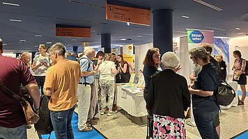 Menschenmenge in einer Messehalle. Mehrere Personen stehen dicht beieinander an einem langen, weißen Tisch mit Informationsmaterialien. Die Personen wirken überwiegend erwachsen bis älter, tragen sommerliche Alltagskleidung, viele mit kurzen Ärmeln, einige mit Rucksäcken oder Umhängetaschen. Im Vordergrund steht eine Person mit dunkelblauem Kleid und schwarzem Rucksack, daneben jemand mit weißem Haar und dunkler Jacke, vor einer Person mit Rock in Rosa-Schwarz-Muster. Entlang des Tisches liegen viele Blätter und Broschüren, teils gelb, teils weiß, dazu einige kleine Aufsteller. Hinter dem Tisch stehen weitere Personen, die offenbar etwas erklären oder zeigen. Über dem Bereich hängen rechteckige Decken-Schilder in Orange mit schwarzer Aufschrift, die einzelne Stände oder Bereiche markieren. Im Hintergrund steht ein Roll Up Banner mit Farbverlauf von Blau zu Grün und der gut lesbaren weißen Aufschrift „besser sehen“. Der Raum hat eine dunkle Decke mit länglichen Lichtleisten und einen hellen Boden.