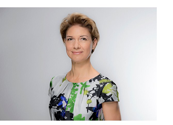 Bild Frau Bürgermeisterin Dr. Alexandra Sußmann. Eine Frau mit kurzen blond-braunen Haaren, geblümtem Oberteil, vor hellgrauem Hintergrund.