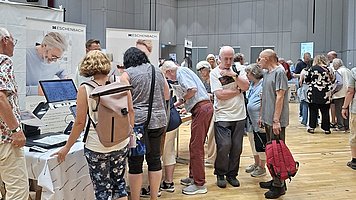Menschenmenge in einer Messehalle. In der Bildmitte stehen viele Personen dicht beieinander in einer Reihe vor einem Stand mit weißen Tischen. Auf den Tischen liegen und stehen mehrere technische Geräte, unter anderem größere Bildschirm-Lupen mit Monitor und Bedienelementen. Einige Personen beugen sich über die Geräte und probieren sie aus oder lassen sich etwas erklären. Links im Hintergrund hängen weiße Roll Up Banner mit dem Aufdruck „ESCHENBACH“ und einem großen Foto eines älteren Mannes mit Brille, der nach unten schaut. Die Besuchenden tragen sommerliche Kleidung: kurze Hosen, T Shirts, Sandalen, Turnschuhe, teils Rucksäcke oder Umhängetaschen. Eine Frau im Vordergrund von hinten trägt einen beigefarbenen Rucksack und eine dreiviertellange gemusterte Leggings. Rechts hält eine Person einen auffälligen roten Rucksack in der Hand. Der Boden besteht aus hellem Holz. Der Raum ist groß und hoch, mit grauen Wänden. Unter der Decke befinden sich mehrere Ebenen von abgehängten Akustikelementen und viele kleine 