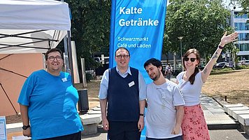 Vier Personen im Freien vor einem blauen Aufsteller. Die Gruppe steht nebeneinander auf einem sonnigen Platz. Links ist ein Mann mit kurzer dunkler Frisur, mittelheller Haut, blauem T Shirt, weiten schwarzen Hosen und leuchtend blauen Sportschuhen, er lächelt. Daneben steht ein Mann mit heller Haut, Brille, blauem ärmellosen Pullover über einem hellblauen Hemd und dunkler Stoffhose, ebenfalls lächelnd. Rechts von ihm steht ein Mann mit kurzer dunkler Frisur, heller Haut, hellblauem T Shirt und blauer Hose, der leicht schräg steht. Ganz rechts steht eine Frau mit heller Haut, langen dunkelblonden bis bräunlichen Haaren, weißem Oberteil und knöchellangem rosa Kleid mit Blumenmuster; sie trägt helle flache Schuhe und streckt fröhlich einen Arm nach oben.  Hinter der Gruppe steht eine große blaue Flag mit dem Text „Kalte Getränke“ und darunter „Schwarzmarkt Café und Laden“. Links im Hintergrund befindet sich ein weißes Pavillon Zelt mit Tisch, rechts und dahinter sind Bäume, Rasenflächen und einige moderne Gebäude zu erkennen.
