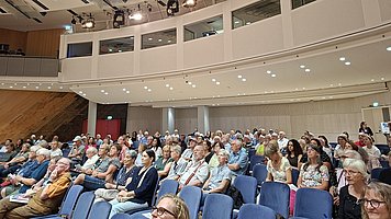 Ein großer Saal mit vielen Zuschauern in Reihen blauer Polstersitze. Der Raum ist gut gefüllt, die Menschen sitzen dicht nebeneinander und wirken überwiegend älter, in sommerlicher Alltagskleidung wie Hemden, Blusen, Kleider und T-Shirts, teils mit Mustern.   Die Sitzreihen verlaufen von unten links nach oben zur Bildmitte, wie in einem ansteigenden Hörsaal oder Theater. Rechts im Bild befindet sich eine hohe, helle Seitenwand mit mehreren rechteckigen Fenstern und integrierten Lautsprechern. An dieser Wand und im Deckenbereich sind zahlreiche runde Deckenleuchten und mehrere große Scheinwerfer montiert. Der Gesamteindruck ist der eines vollen, modernen Veranstaltungs- oder Konferenzsaals kurz vor Beginn oder während einer Veranstaltung.