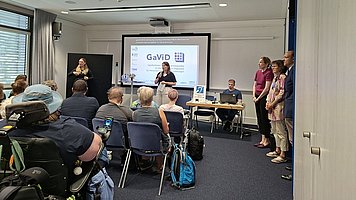 Ein Vortragsraum mit vielen sitzenden Zuhörerinnen und Zuhörern, die auf eine Leinwand und eine Referentin vorne im Raum ausgerichtet sind. Vorne steht eine Frau im hellen Blazer mit Namensschild und spricht zu dem Publikum, einige Personen sitzen in Rollstühlen. Auf der Leinwand ist eine Präsentationsfolie projiziert. Darauf steht (größter Text in der Mitte): „‚Schlecht sehen – selbstständig bleiben‘“ Darunter: „Wohnanpassung bei Sehbeeinträchtigung. Sabine Backmund – BSV Württemberg e.V.“ Am unteren Rand der Folie steht: „Mit freundlicher Unterstützung“ Links unten auf der Folie ist das Logo des „Blinden- und Sehbehindertenverband Württemberg e.V.“ zu erkennen, rechts unten das Logo von „Aktion Mensch“.