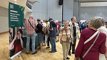 Menschenmenge in einem Veranstaltungssaal.  Auf dem Foto stehen viele Personen auf einem hellen Parkettboden in einem großen, modernen Saal mit hoher Decke und grauen Wandpanelen. Im Vordergrund ist eine Person mit kurz geschnittenen grauen Haaren, pinkfarbenem langen Oberteil und einer diagonalen, hellen Umhängetasche von hinten zu sehen. Davor stehen weitere Personen, die sich unterhalten; die meisten tragen Freizeitkleidung wie Jeans, Hemden und T Shirts, einige haben Rucksäcke oder Kameras umgehängt.  Links im Bild steht ein großes, senkrechtes Werbebanner mit grünem oberem Teil. Darauf ist oben ein Logo mit weißen Linien zu erkennen, darunter in Weiß der Text: „Besser sehen. Beratung Hilfsmittel Schulungen“.  Unter dem Text ist ein großes Foto auf dem Banner: Es zeigt eine Person mit kurzen dunklen Haaren, rotem Oberteil und schwarzem Schal, die eine Brille aufsetzt. Vor dem Banner befindet sich ein Tisch mit grüner Tischdecke, an dem mehrere Personen stehen. Der Saal ist hell beleuchtet, bei der Decke ist eine geschwungene, hellbraune Konstruktion mit eingelassenen Spots zu erkennen. Im Hintergrund stehen weitere Gruppen von Menschen.