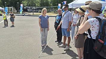 Gruppe von Menschen, die nebeneinander auf einem breiten, sonnigen Platz stehen. Im Vordergrund stehen mehrere Erwachsene in einer Reihe, seitlich zur Kamera gedreht. Die vorderste Person trägt einen hellen Hut, ein helles T Shirt mit kleinen roten Mustern und einen großen, grau blauen Rucksack. Daneben und dahinter stehen weitere Personen mit Sommerkleidung wie T Shirts, kurze Hosen und Hüte, einige mit Rucksäcken oder Umhängetaschen. Viele tragen Sonnenhüte oder Kappen und Sonnenbrillen. Etwas vor der Gruppe, links im Bild, steht eine Frau mit heller Haut, kurzem blonden Haar, grauem T Shirt und grauer Hose. Sie hält einen langen weißen Langstock schräg nach vorn auf den Boden gerichtet. Im Hintergrund verläuft die breite Asphaltstraße mit aufgemalten weißen Linien. Weiter hinten stehen Menschen mit gelben Warnwesten und eine Person mit dunkler Kleidung, alle auf der Straße verteilt. Am Rand sind Pavillons, Fahnen und Banner sowie Bäume zu erkennen.