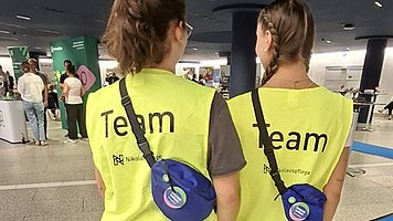 Zwei Frauen stehen mit dem Rücken zur Kamera in einer großen Halle. Beide tragen gelbe, ärmellose Warnwesten mit schwarzer Aufschrift „Team“ oben auf dem Rücken. Beide haben eine kleine, runde, dunkelblaue Umhängetasche auf dem Rücken, befestigt mit einem schwarzen Gurt. Die Person links trägt ein dunkelbraunes T Shirt, eine hellblaue Jeans und weiße Turnschuhe. Die Person rechts trägt ein kurzärmeliges Oberteil, eine graue enge Hose und ebenfalls weiße Turnschuhe.  Sie gehen nebeneinander auf einem langen, leuchtend blauen Läuferteppich, der sich durch die helle, körnig gemusterte Bodenfläche zieht.  Im Hintergrund stehen mehrere Menschen an Tischen; darüber befinden sich dunkle Deckenpaneele mit vielen kleinen Lichtern.