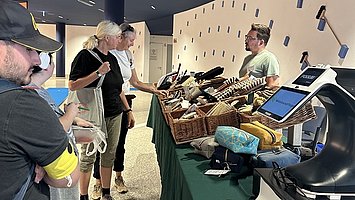 Ein langer Tisch mit grüner Tischdecke in einem Innenraum.   Auf dem Tisch stehen mehrere geflochtene Körbe voll mit verschiedenen Besen und Bürsten in unterschiedlichen Größen und Formen. Hinter dem Tisch steht rechts ein Mann mit kurzem Haar und Brille, vor ihm ein Kassenterminal, das blau leuchtet. Links daneben, vor dem Tresentisch, stehen zwei Frauen: eine mit hellblonden, zum Zopf gebundenen Haaren in einem weißen Oberteil, die andere mit etwas dunkleren Haaren in einem dunklen Oberteil; beide sind dem Tisch zugewandt und betrachten die ausgelegten Produkte.   Ganz links stehen weitere Personen dicht beieinander; eine Person trägt eine dunkle Kappe, ein graues T Shirt und auffällig gelbe Shorts sowie eine Tasche, eine andere Person dahinter trägt ein dunkles T Shirt und hellere, knielange Hosen. Der Hintergrund besteht aus einer hellen Wand mit vielen kleinen, rechteckigen, dunkelblauen Dekoteilen, darüber eine dunklere Decke mit vereinzelten Leuchtstreifen.
