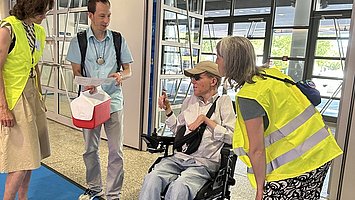 Messeguides in gelben Warnwesten unterstützen einen Besucher im Rollstuhl.