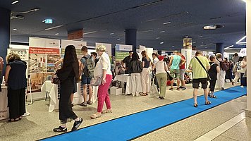 Messestand in einer Messehalle mit vielen Menschen. Mehrere Stände mit Roll ups und großen Plakaten sind in einer breiten Reihe angeordnet, davor stehen und gehen viele Besucherinnen und Besucher. Mittig verläuft ein schmaler, kräftig blauer Teppichstreifen als Wegführung durch die Halle. Links des Teppichs liegt ein heller Boden, rechts davon stehen die einzelnen Stände mit weißen Tischen und teilweise weißen Hussen über den Stühlen. Direkt im Vordergrund sind mehrere Personen im Freizeit  oder Sommeroutfit zu sehen, etwa eine Person mit neongelbem Oberteil und schwarzer Hose sowie eine Person mit pinkfarbener Hose. Weiter hinten unterhalten sich Gruppen vor den Ständen, einige schauen auf Informationsmaterial, andere sprechen mit Standpersonal.   Über den Ständen hängen große Plakate und Banner mit Text und Bildern, die verschiedene Angebote oder Firmen bewerben. Die Decke der Halle ist dunkel, mit länglichen, hellen Lichtstreifen und runden Einbauten, was der Szene ein modernes, leicht gedämpftes Licht gibt. Im Hintergrund ist ein leuchtendes, blaues Notausgangsschild zu erkennen.