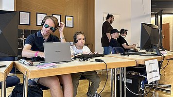 Eine Szene in einem Raum mit mehreren Personen, die an Tischen mit Computern sitzen und Kopfhörern tragen. Im Vordergrund sitzt links ein erwachsener Mann mit kurzen blonden Haaren, Sonnenbrille und großen schwarzen Kopfhörern vor einem silberfarbenen Laptop. Er trägt ein dunkles Polohemd, kurze Hosen, blaue Socken und dunkle Sportschuhe. Neben ihm sitzt ein junger Mann mit lockigen, mittelblonden Haaren und großen schwarzen Kopfhörern, er schaut konzentriert auf einen Laptop vor sich. Auf dem Tisch liegen verschiedene Kabel, ein kleines Audio Mischpult, eine Computermaus und ein paar Zettel. Unter dem Tisch stehen eine schwarze Rucksacktasche und eine weiße Mehrfachsteckdose mit mehreren eingesteckten Netzteilen und Kabeln, die mit Klebeband am hellen Holzboden gesichert sind. Weiter hinten im Raum sitzen rechts noch zwei Personen mit Kopfhörern an einem großen Monitor; hinter ihnen steht eine weitere Person und beobachtet sie. An einem der Tische hängt ein Schild, auf dem groß „Gaming“ steht. Der Raum wirkt hell, mit Holzboden und einer Wandnische aus Holz, in der mehrere gerahmte Bilder hängen.